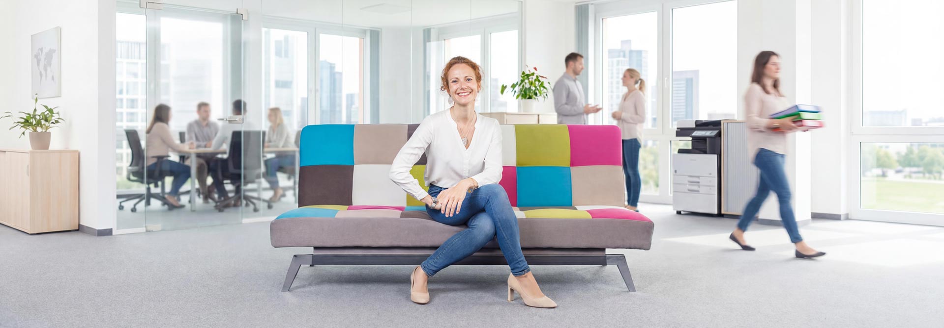 Eine Frau sitzt in einer modernen Bürolounge, während im Hintergrund eine weitere Person durch den Raum geht.