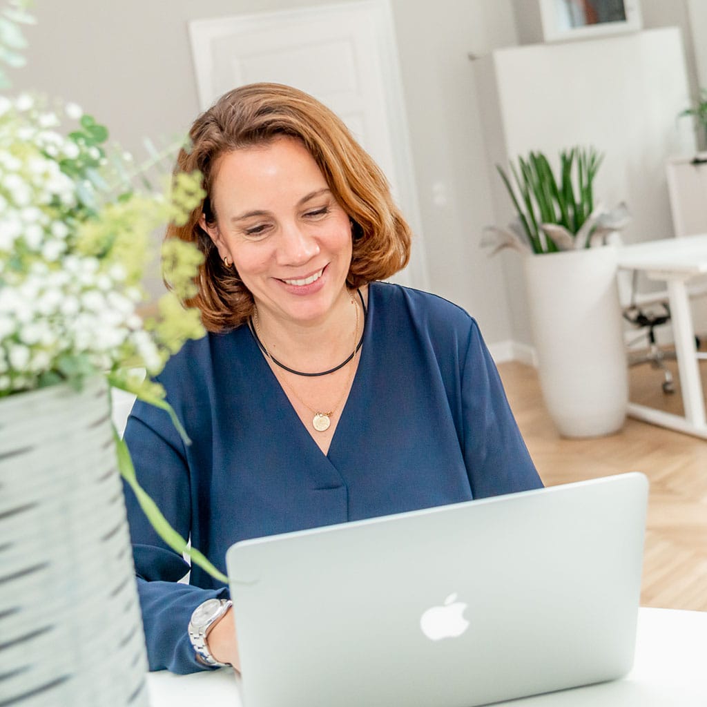 Frau sitzt lächelnd an einem Laptop in einem hellen modernen Büro.