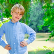 Kindergartenfotograf in Fürth macht ein Foto, auf dem ein blonder Junge in weißer Hose und blauem Hemd, unter den Bäumen im Kindergarten, gezeigt wird.