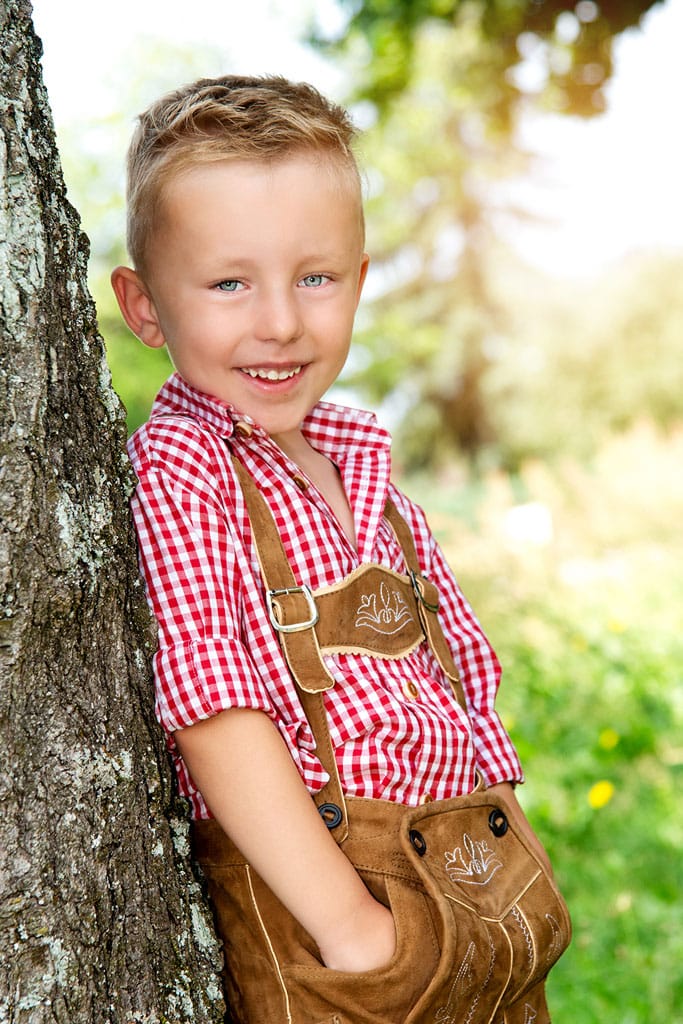 Fotoshooting im Kindergarten mit einem Jungen, der eine bayerische Tracht trägt und an einem Baum lehnt.