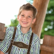 Fröhlicher Junge hat sich mit blauem Hemd und Lederhose für das Kindergarten Fotoshooting im Garten schick gemacht.