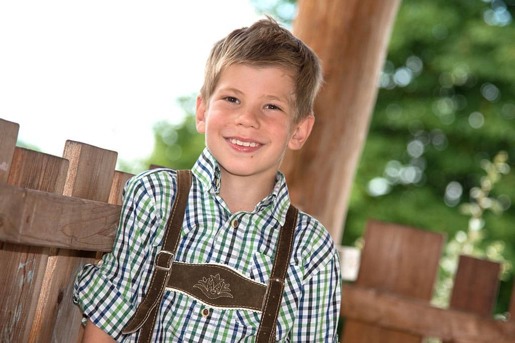 Fröhlicher Junge hat sich mit blauem Hemd und Lederhose für das Kindergarten Fotoshooting im Garten schick gemacht.