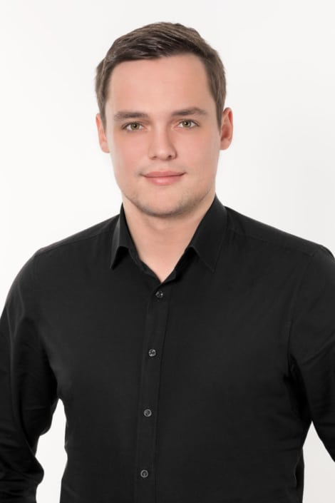 Porträt eines jungen Mannes in einem schwarzen Hemd mit kurzen dunklen Haaren und freundlichem Gesichtsausdruck vor hellem Hintergrund.