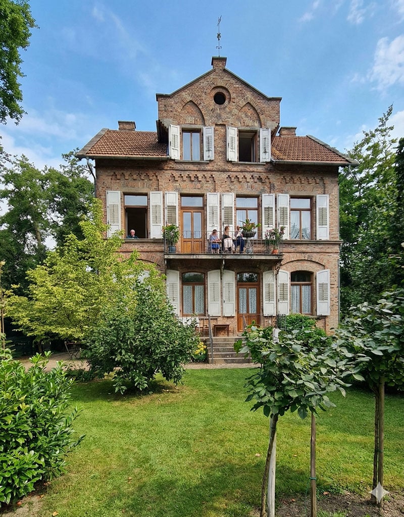 Wenn KI ein altes Foto nicht restauriert, sondern ersetzt Modern wirkende farbige Darstellung eines alten Gebäudes mit Garten, erzeugt aus einer historischen Fotovorlage durch KI.
