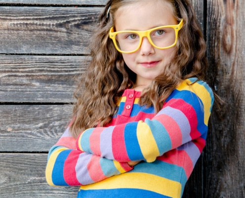 Kindergartenkind mit gelber Brille und bunten Pullover lehnt am Zaun