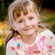 Lachendes blondes Mädchen, mit Zöpfen und weiß/rosa Pullover, lässt sich im Kindergarten fotografieren.
