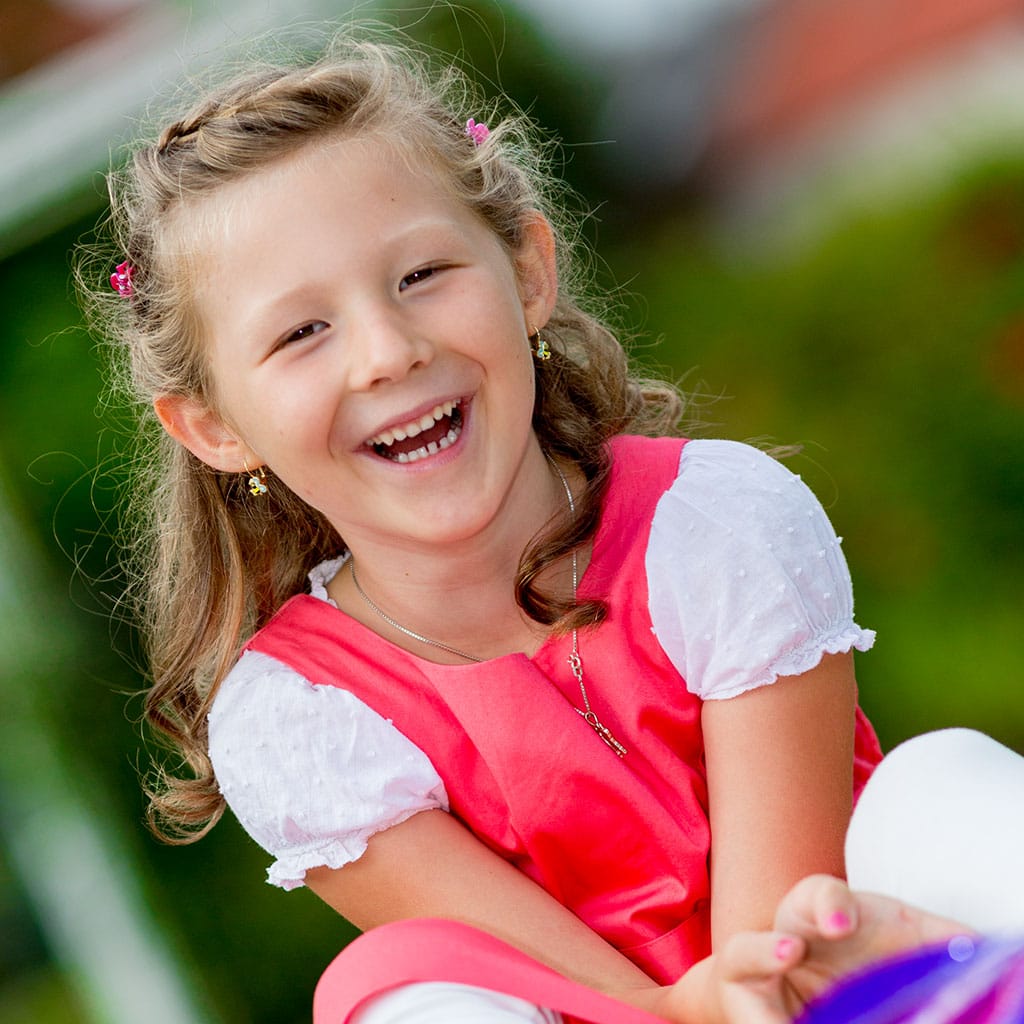 Blondes kleines Mädchen mit rotem Sommerkleid, sitzt lachend auf einem Bobbycar, das für Fotos im Kindergarten zur Verfügung gestellt wurde.