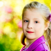 Blondes Kindergartenmädchen im lila Mantel nimmt am Kindergarten Fotoshooting teil und blickt in die Fotokamera. Hintergrund mit schönem Bokeh