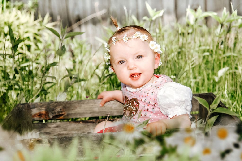 Süßes Kinderfoto, im Kinderhort aufgenommen. Lachendes Baby sitzt mit Blumenkranz im Haar in der Wiese