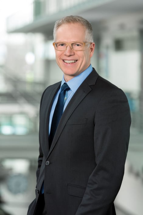 Professionelles Business-Foto eines lächelnden Managers mit Brille im dunklen Anzug vor einem modernem, hellen Büro-Hintergrund.