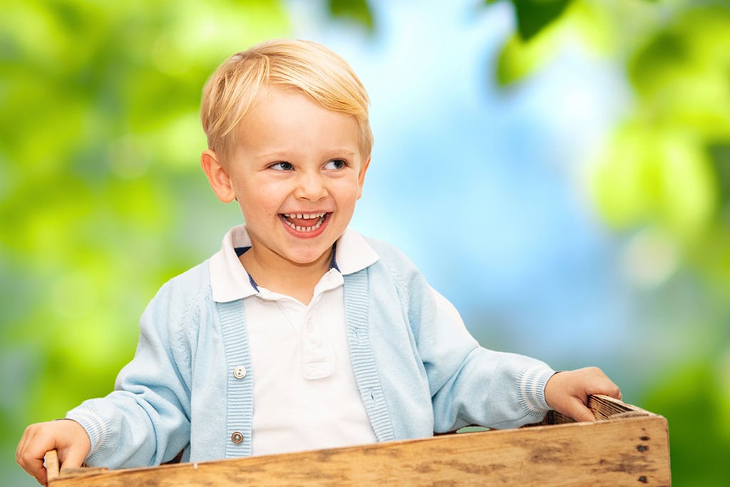 Blondhaariger Junge sitzt in einer Holzkiste im Kindergarten, bereit für coole Kindergartenfotos.