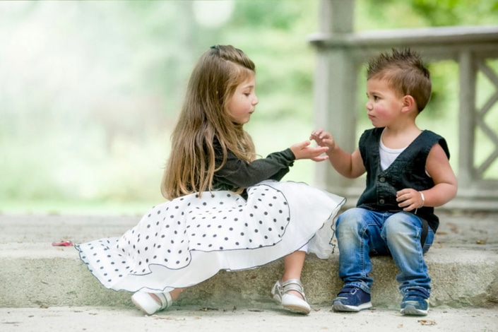 Kindergartenfotografie einmal anders. Schick angezogenes Geschwisterpaar sitzt im Kindergarten auf einer Steinmauer und wird fotografiert.