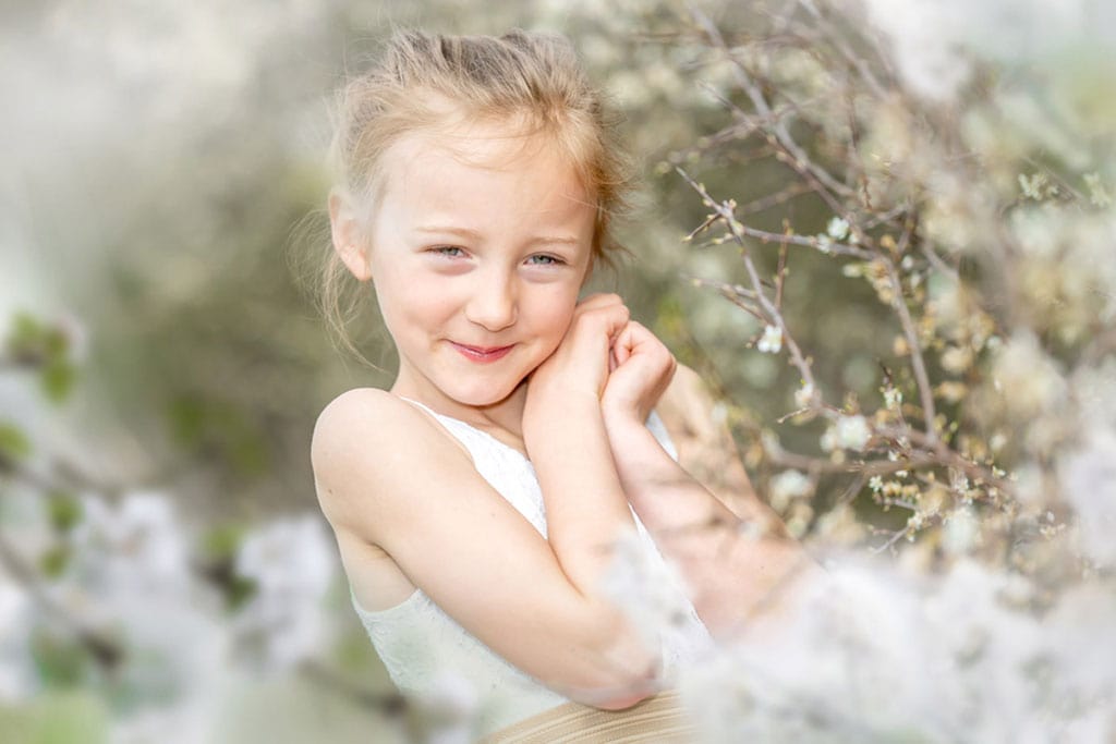 Niedliches Kindergartenfoto, leicht entsättigt. Lächelndes Kind im weißen Kleid sitzt im Blumenmeer.